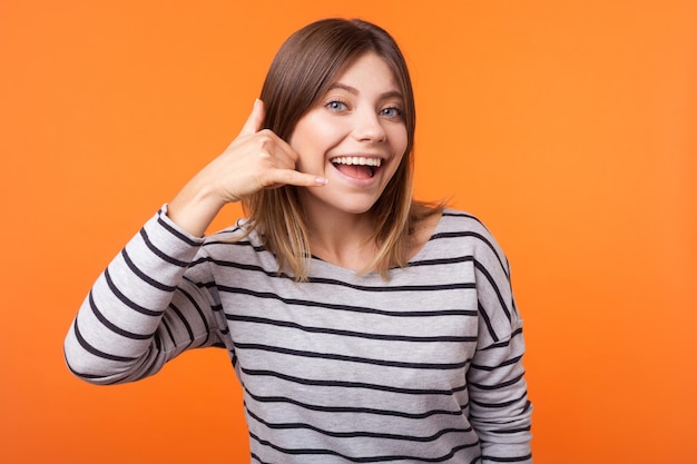 Chame-me Retrato de uma mulher bonita alegre com cabelo castanho na camisa listrada de manga comprida em pé fazendo gesto de chamada com a mão olhando divertido estúdio interior tiro isolado em fundo laranja