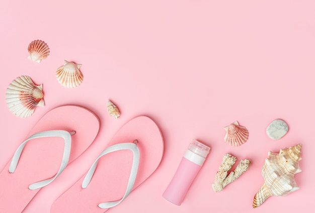 Chanclas, botella de protector solar y conchas sobre fondo rosa pastel, espacio de copia, endecha plana.
