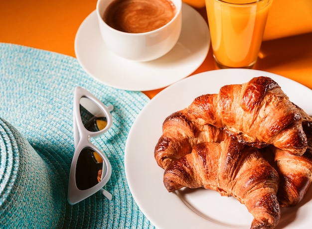 Chapéu, croissant de óculos de sol com uma xícara de café e um copo de suco de laranja em uma mesa amarela ao sol