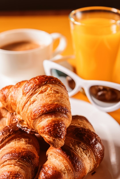 Foto chapéu, croissant de óculos de sol com uma xícara de café e um copo de suco de laranja em uma mesa amarela ao sol