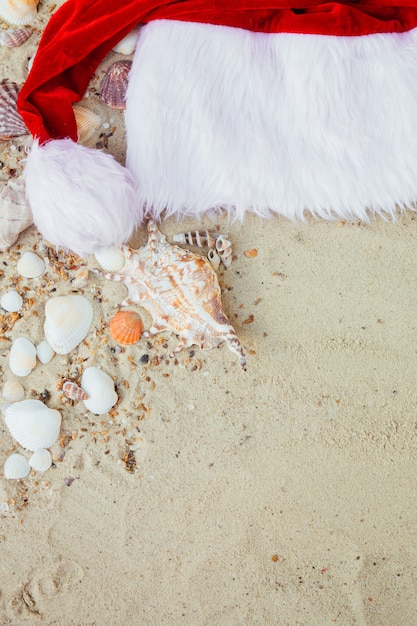 Chapéu de Natal na praia. Chapéu de Papai Noel na areia perto de conchas.