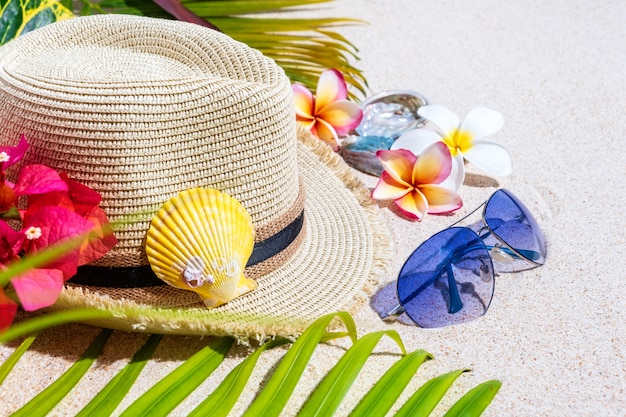 Chapéu de palha bege com óculos de sol azuis, conchas do mar colorido, flores de frangipani e folhas de palmeira verde na areia.