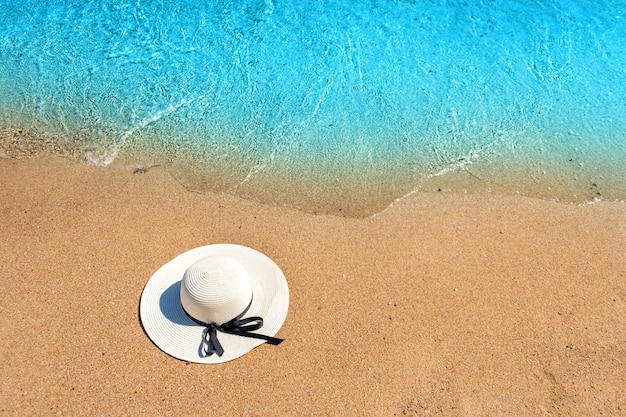 Chapéu de palha da mulher branca que coloca na praia tropical da areia com água vibrante azul do oceano no fundo no dia de verão ensolarado. Férias e conceito de viagens de destino.