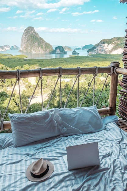 chapéu, laptop e travesseiro em uma cama em uma varanda de madeira com vista para o mar e as montanhas