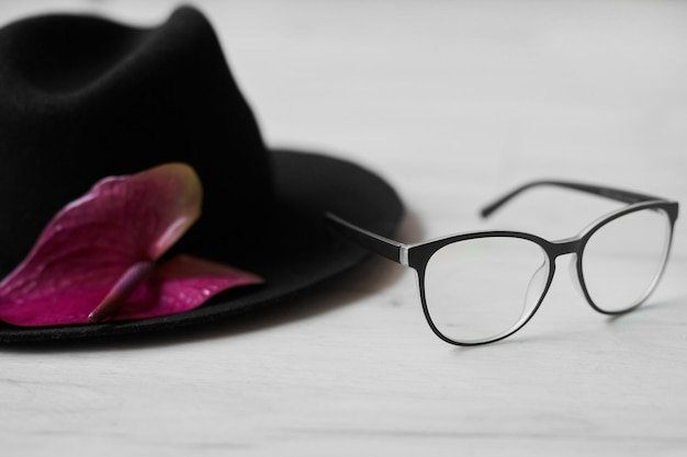 Chapéu preto elegante com uma flor rosa fica ao lado de óculos da moda em uma mesa de madeira branca