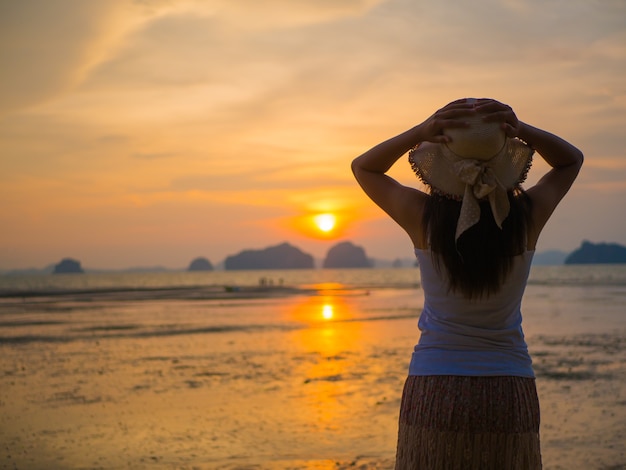 Chapéu vestindo da mulher com os braços abertos sob o nascer do sol perto do mar.