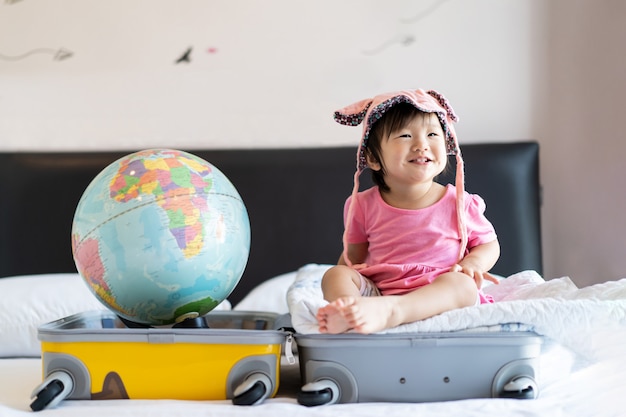 Chapéu vestindo do bebê pequeno bonito asiático que senta-se na mala de viagem com o sorriso que sente engraçado e que ri na cama no quarto.
