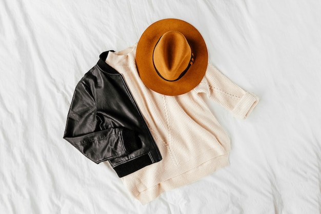 Foto chaqueta de cuero y suéter blanco cálido con sombrero marrón sobre sábana blanca. traje elegante de otoño o invierno para mujer. collage de ropa de moda. vista plana endecha, superior.