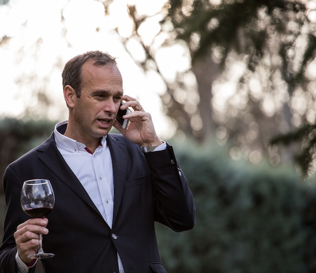 Foto chaqueta hombre habla por teléfono mientras bebe una copa de vino en el campo