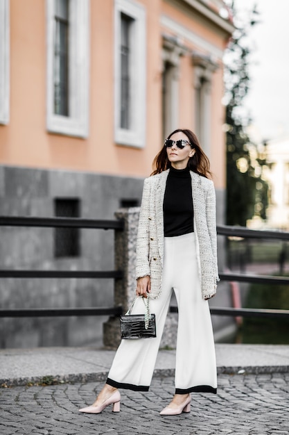 Chaqueta morena chica de moda y pantalón blanco caminando por la ciudad con un bolso negro y gafas de sol.