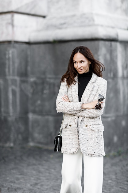 Chaqueta morena chica de moda y pantalón blanco caminando por la ciudad con un bolso negro y gafas de sol.