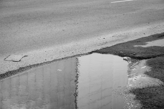 Charco en carretera en escala de grises. La pared del edificio reflejada en el agua en tiempo lluvioso se cierra para arriba. Apatía, depresión, tristeza en Rusia. Basura en el asfalto.