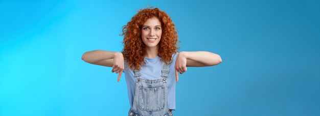 Foto charismatische, freche, kokette rothaarige, gewagtes, rothaariges mädchen, lockiger haarschnitt, der mit dem zeigefinger nach unten zeigt