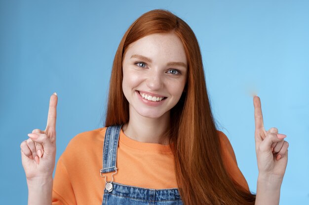 Charismatische gut aussehende europäische rothaarige Frau der Taille, die aufrichtig lächelt und gerne zeigt, wie Zeigefinger nach oben zeigen glückliche Hilfe empfehlen coolen Werbespot blauen Hintergrund