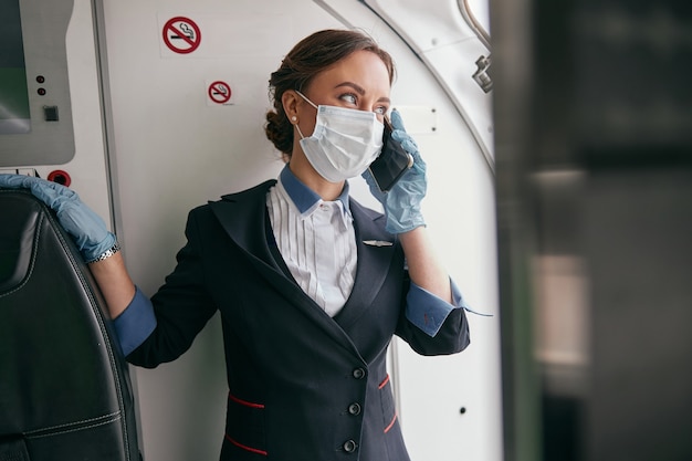 Charla de azafata por teléfono móvil en el avión de pasajeros. Interior del avión moderno. La mujer europea usa uniforme, guantes de látex y máscara médica. Aviación civil comercial. Concepto de viaje aéreo