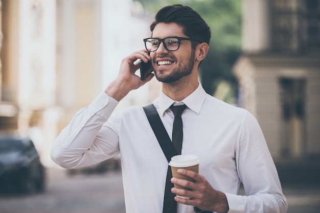 Charla de negocios sobre la marcha. Joven alegre en vasos sosteniendo una taza de café y hablando por teléfono móvil con una sonrisa mientras camina al aire libre