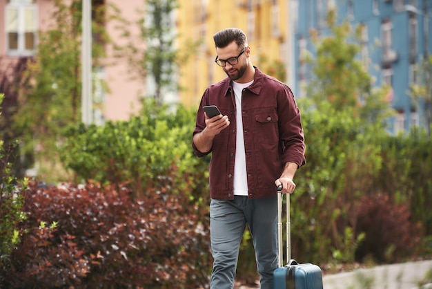 Charlando con un amigo. Hombre guapo con barba en ropa casual y anteojos tirando de su equipaje y usando el teléfono inteligente mientras camina por la calle de la ciudad. Concepto de viaje. Estilo de vida