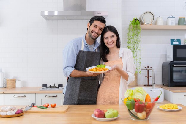 Charmant aussehendes junges Paar, das sanft in die Kamera blickt, die ein Valentinstag-Ernährungsereignis vorbereitet Frau mit Schüsselsalat-Makkaroni-Hersteller zeigt Romantik in der heimischen Küche