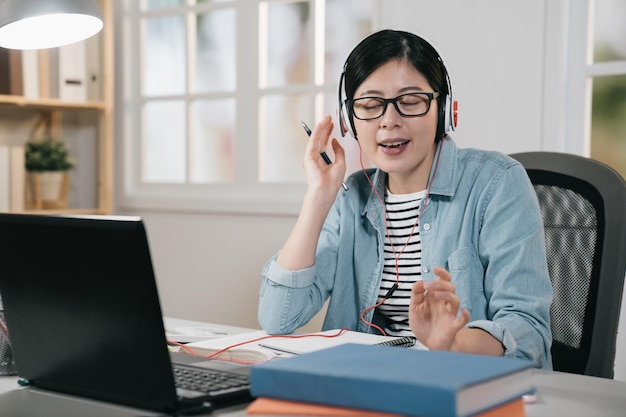 charmante asiatische chinesische Teenager-Studentin mit Laptop-Computer und Kopfhörern auf dem Desktop. Entspannen Sie sich, schließen Sie die Augen der College-Frau, genießen Sie Musik im Headset und singen Sie, während Sie tagsüber zu Hause lernen.