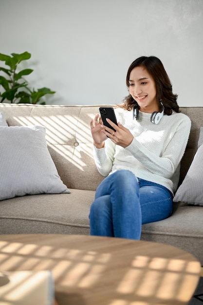 Charmante asiatische Frau, die ihr Smartphone benutzt, während sie sich auf ihrer Couch im Wohnzimmer ausruht
