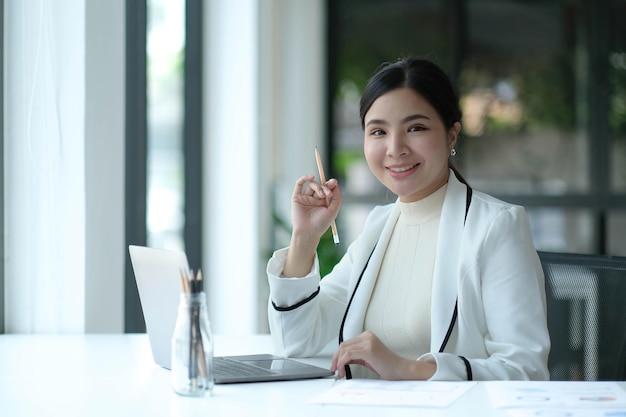 Charmante asiatische Frau, die mit einem Laptop im Büro arbeitet und in die Kamera schaut