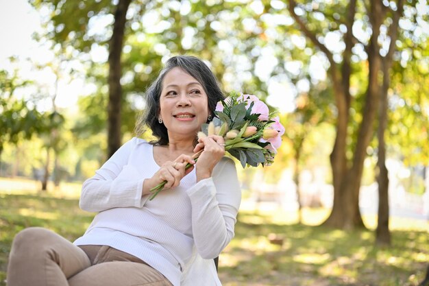 Charmante asiatische Frau im Alter der 60er Jahre, die einen schönen Blumenstrauß hält, während sie sich im Park entspannt