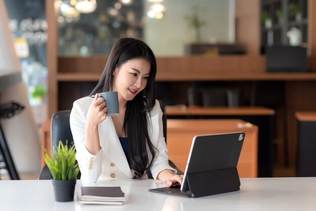 Charmante asiatische Geschäftsfrau, die eine Kaffeetasse hält und an einem Tablet im Büro arbeitet.