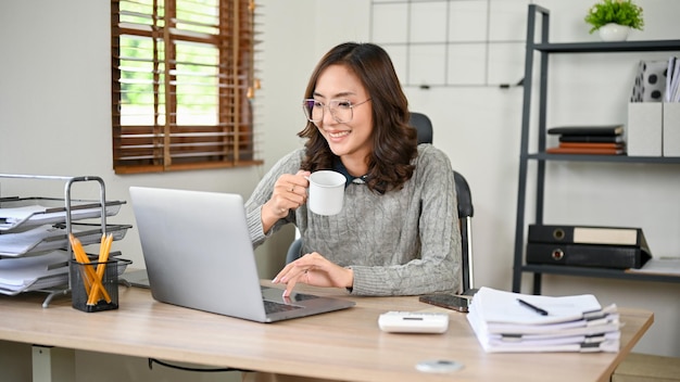 Charmante asiatische Geschäftsfrau, die Kaffee trinkt, während sie an ihren Aufgaben auf dem Laptop arbeitet