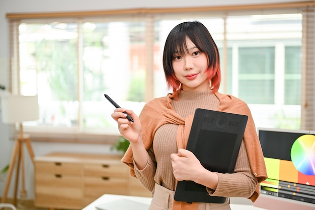 Charmante asiatische Grafikdesignerin steht in ihrem Büro und hält Stift und Grafiktablett