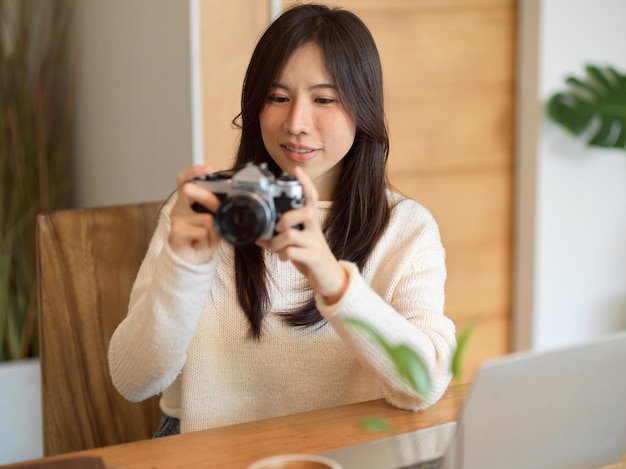 Charmante asiatische junge Fotografin, Inhaltsredakteurin mit Vintage-Retro-Kamera, sitzt und arbeitet an ihrem Laptop im Café.