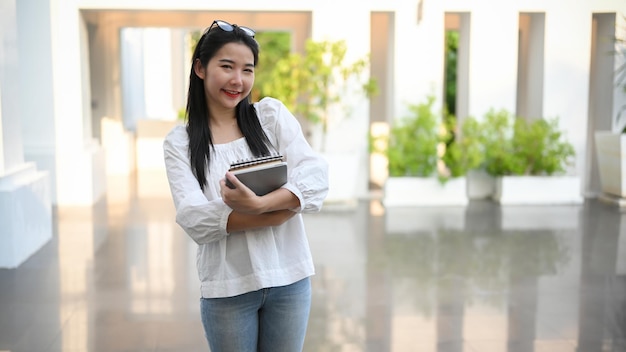 Charmante asiatische Studentin, die vor dem Campusgebäude steht und ihr Lehrbuch hält