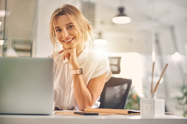 Charmante blonde Dame, die lächelt, während sie mit Laptop bei der Arbeit am Tisch sitzt