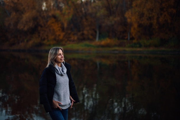 Charmante blonde Frau, die am Fluss steht und wegschaut, lächelnd mit schwarzer Jacke auf den Schultern, Händen auf Pullover mit Bäumen, die mit Sonnenuntergang im Hintergrund bedeckt sind Herbstspaziergang im Wald