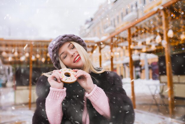 Charmante blonde Frau, die auf dem mit Lichtern geschmückten Weihnachtsmarkt leckere Ingwerkekse isst