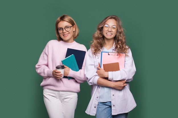 Charmante blonde Studenten lächeln auf einer grünen Wand, während sie einige Bücher umarmen und einen Kaffee trinken