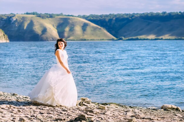 Charmante Braut im weißen Kleid, die am Ufer eines Sees geht