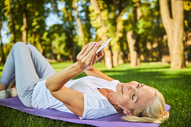 Charmante Dame, die eine SMS auf dem Smartphone schreibt und lächelt, während sie auf grünem Gras liegt