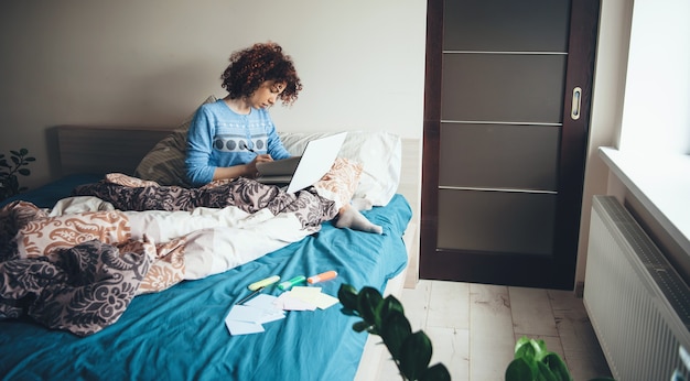 Charmante Dame mit lockigem Haar, die einen blauen Pyjama trägt, benutzt einen Laptop im Bett, während sie Hausaufgaben macht