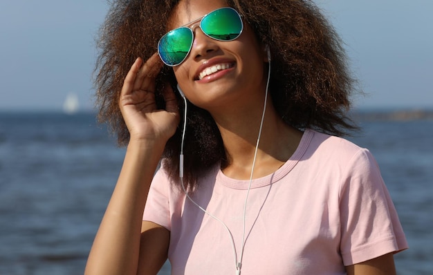 Charmante, erstaunliche afroamerikanische junge Frau mit Sonnenbrille, die Musik in Kopfhörern auf ihrem Handy hört Lässig gekleidet