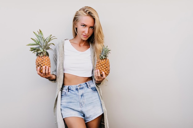 Charmante europäische Frau in Denim-Shorts mit exotischen Früchten Innenfoto eines sinnlichen kaukasischen Mädchens mit Ananas