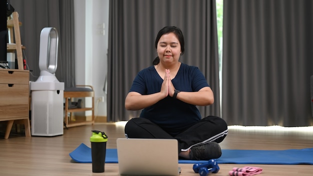 Charmante fettleibige Frau praktiziert Yoga mit Laptop zu Hause.