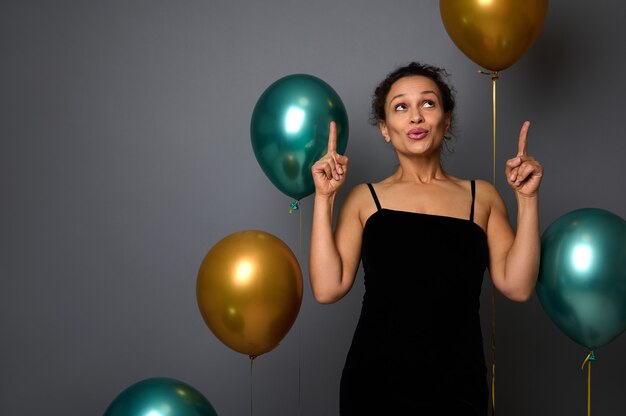 Charmante Frau, die ein schwarzes Samtabendkleid trägt, lächelt und zeigt mit den Zeigefingern auf einen Kopienraum auf grauem Hintergrund mit Luftballons. Geburtstag, Weihnachten, Neujahr Konzept