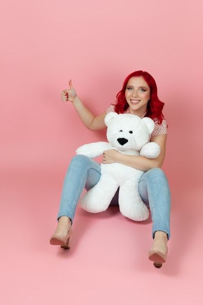 charmante Frau in Jeans mit roten Haaren hält einen großen weißen Teddybär und gibt einen Daumen hoch