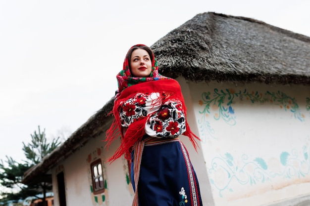 Charmante Frau in traditioneller ukrainischer Taschentuchhalskette und besticktem Kleid, die im Hintergrund einer dekorierten Hütte im ukrainischen Stil steht