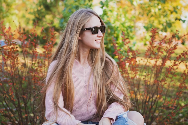 Charmante Frau mit langen Haaren und in lässiger Sonnenbrille sitzt auf der Bank im schönen Herbstpark