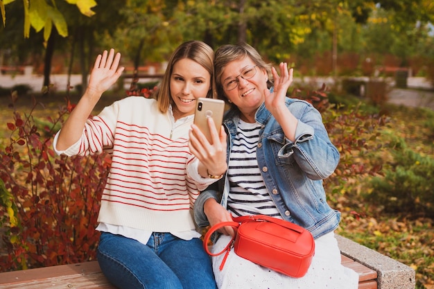 Charmante Frauen im lässigen Stil Mutter und Tochter sitzen auf einer Bank in einem wunderschönen Herbstpark und Video-Chat auf dem Smartphone