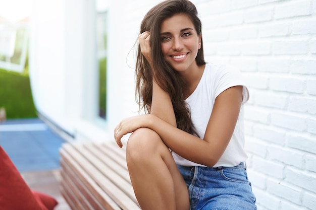 Charmante Freundin sitzt auf einer Außenbank in der Nähe einer weißen Backsteinmauer