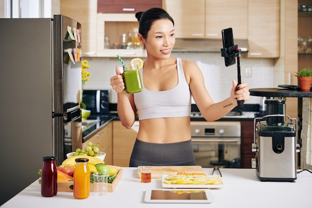 Charmante junge Asiatin, die sich selbst beim Trinken eines gesunden grünen Smoothies aufzeichnet