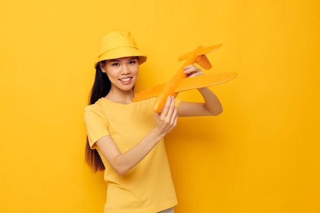 Charmante junge asiatische Frau in einem Hut mit einem Flugzeug in seinen Händen modellieren den gelben Hintergrund des Spielzeugs unverändert