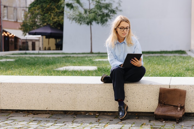 Charmante junge Blondine in Brillen, die sich an der frischen Luft entspannen und ein digitales Tablet verwenden. Konzept der modernen Technologie und des urbanen Lebensstils.